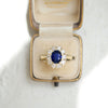 A woman wearing a blue sapphire and diamond cluster ring in a vintage velvet ring box.